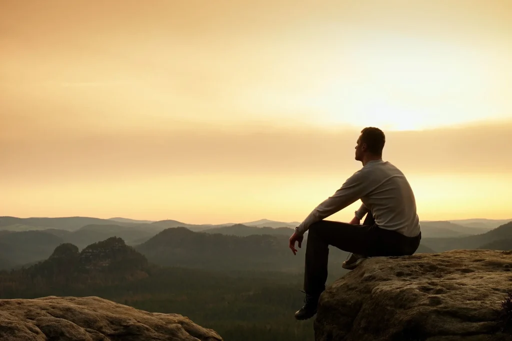 Man sitting on top of a mountain looking up at the sky in contemplation.