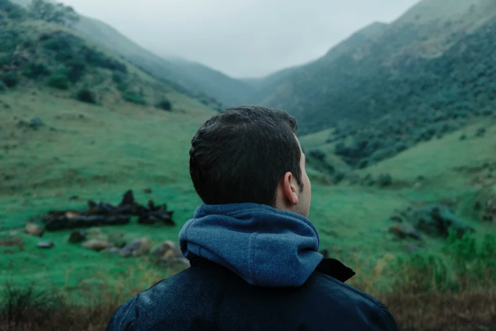 Person standing with their back to the camera, looking out over a green mountain valley with rolling hills and cloudy sky.