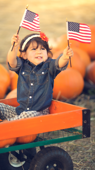 Toddler on a trolley with a flag in each hand and pumpkins in the background.